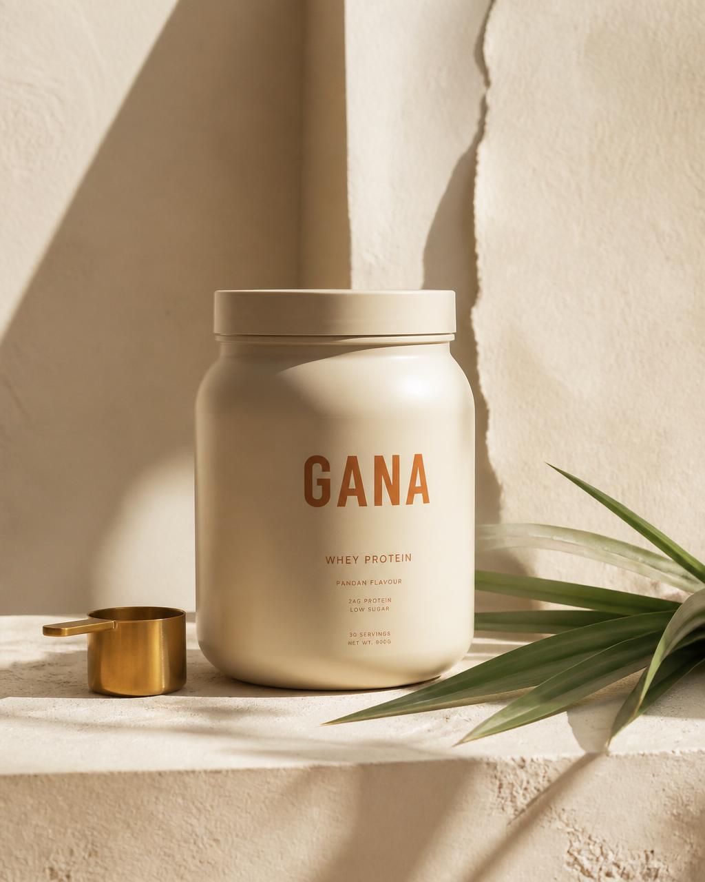 GANA protein tub with brass scoop and pandan leaf on warm cream surface